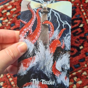 May include: A tarot card depicting a lighthouse being attacked by a giant octopus in a stormy sea. The card is labeled "The Tower" in white script. The image is set against a red and blue patterned background.