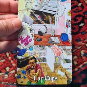 May include: A tarot card illustration depicting a chaotic kitchen scene with floating objects and a woman holding a child. The card is labeled "4 of Cups" and features a mug with the text "Live Laugh Love".
