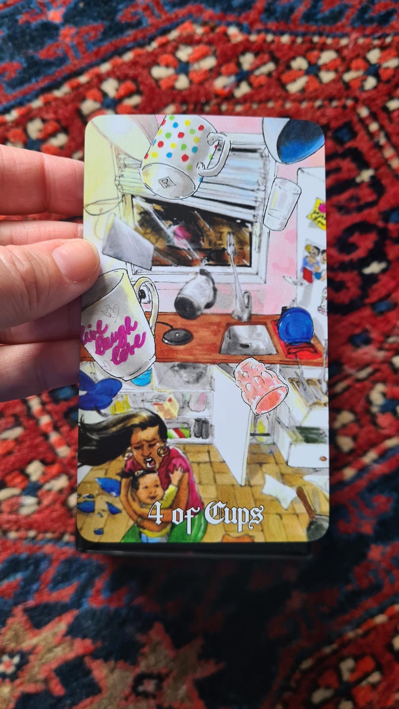 May include: A tarot card illustration depicting a chaotic kitchen scene with floating objects and a woman holding a child. The card is labeled "4 of Cups" and features a mug with the text "Live Laugh Love".