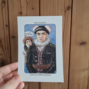 May include: A cartoon illustration of a character from the British comedy series Blackadder. The character is wearing a black jacket, a white collar, and a red and black patterned vest. He has a surprised expression on his face and is holding a pink pig ear in each hand. The text "We got a cunning plan!" is written in a speech bubble above his head. The text "Blackadder" is at the top of the card and "Edmund Blackadder" is at the bottom.