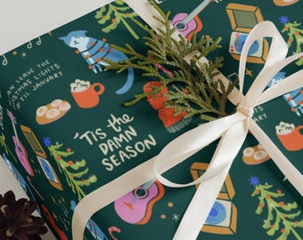 May include: Dark green wrapping paper featuring a repeating pattern of guitars, cats wrapped in Christmas lights, mugs of cocoa, and Christmas trees. The text reads 'Tis the Damn Season' and 'I can leave the Christmas lights up til January'.