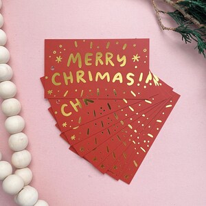 May include: A stack of red gift tags with gold foil lettering that reads "*MERRY* CHRISTMAS!" The tags are decorated with gold foil stars and dots.
