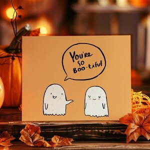 Op de afbeelding: Een Halloween-kaart met een griezelige boodschap die "You're so Boo-tiful" luidt. De kaart toont twee vriendelijke geesten met glimlachende gezichten. De kaart ligt op een tafel met een verlichte jack-o-lantern en herfstbladeren.