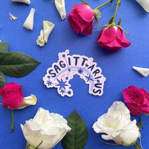 May include: A white sticker with the text "Sagittarius" in a blue and pink arc with stars and a blue starburst. The sticker is on a blue background with white rose petals and pink roses.