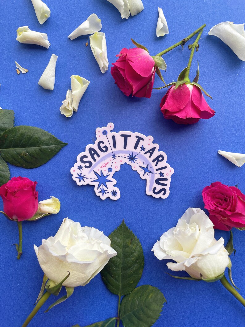 May include: A white sticker with the text "Sagittarius" in a blue and pink arc with stars and a blue starburst. The sticker is on a blue background with white rose petals and pink roses.