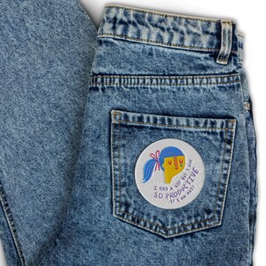 K&ouml;nnte beinhalten: Eine blaue Jeanshose mit einem Patch auf der Ges&auml;&szlig;tasche. Der Patch ist wei&szlig; mit einer Comiczeichnung einer Person mit blauen Haaren und gelber Haut. Der Text auf dem Patch lautet "Ich weine viel, aber ich bin so produktiv, es ist eine Kunst".