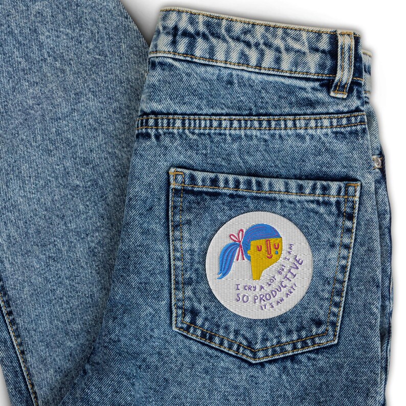 K&ouml;nnte beinhalten: Eine blaue Jeanshose mit einem Patch auf der Ges&auml;&szlig;tasche. Der Patch ist wei&szlig; mit einer Comiczeichnung einer Person mit blauen Haaren und gelber Haut. Der Text auf dem Patch lautet "Ich weine viel, aber ich bin so produktiv, es ist eine Kunst".