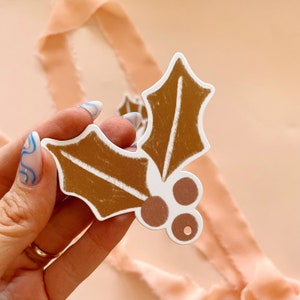 May include: A white paper holly leaf ornament with brown painted leaves and pink painted berries. The ornament has a hole punched in the top for hanging.