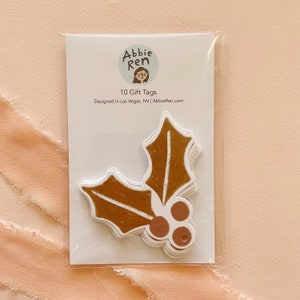 May include: A package of 10 brown and pink holly leaf gift tags with a hole for string. The package has a white background with the text "Abbie Ren" and "10 Gift Tags" in black text. The text "Designed in Las Vegas, NV | AbbieRen.com" is also printed on the package.