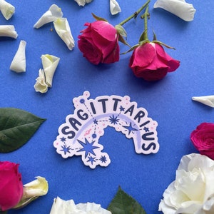 May include: A white sticker with the word "Sagittarius" written in a rainbow arc with stars and constellations. The sticker is on a blue background with white rose petals and red roses.