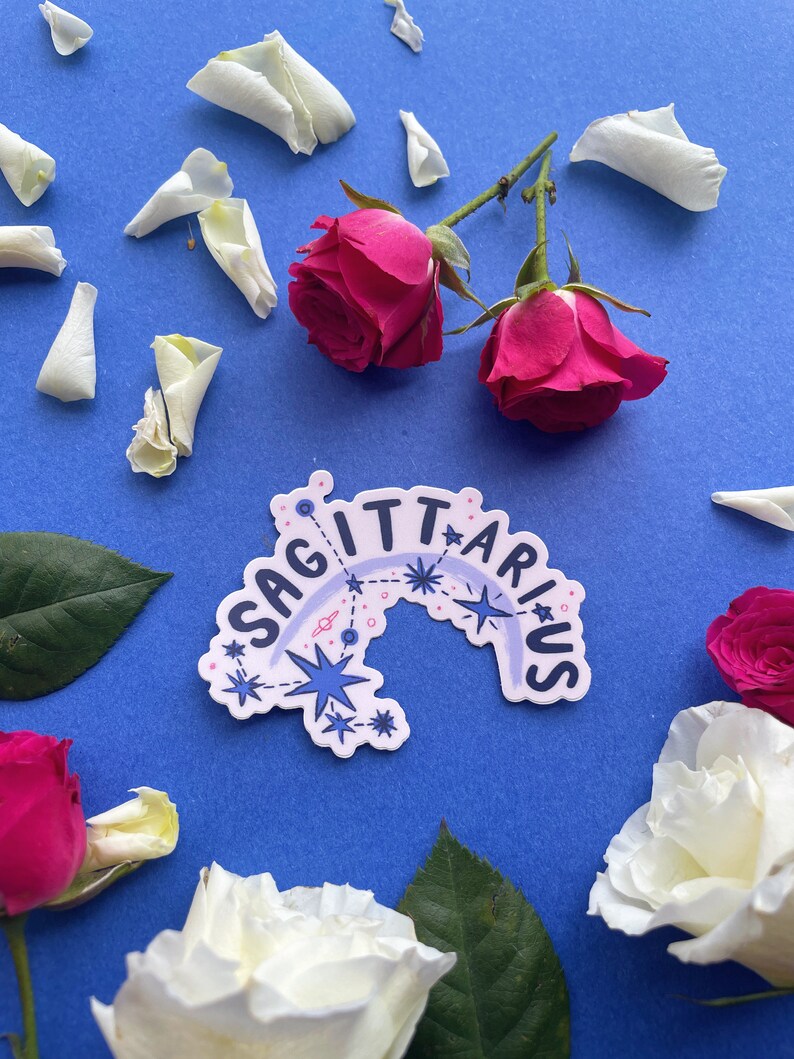 May include: A white sticker with the word "Sagittarius" written in a rainbow arc with stars and constellations. The sticker is on a blue background with white rose petals and red roses.