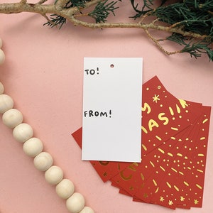 May include: A white gift tag with the words "TO!" and "FROM!" written in black marker. The tag is sitting on a pink surface with a stack of red gift tags with gold foil lettering that says "Merry Christmas!"