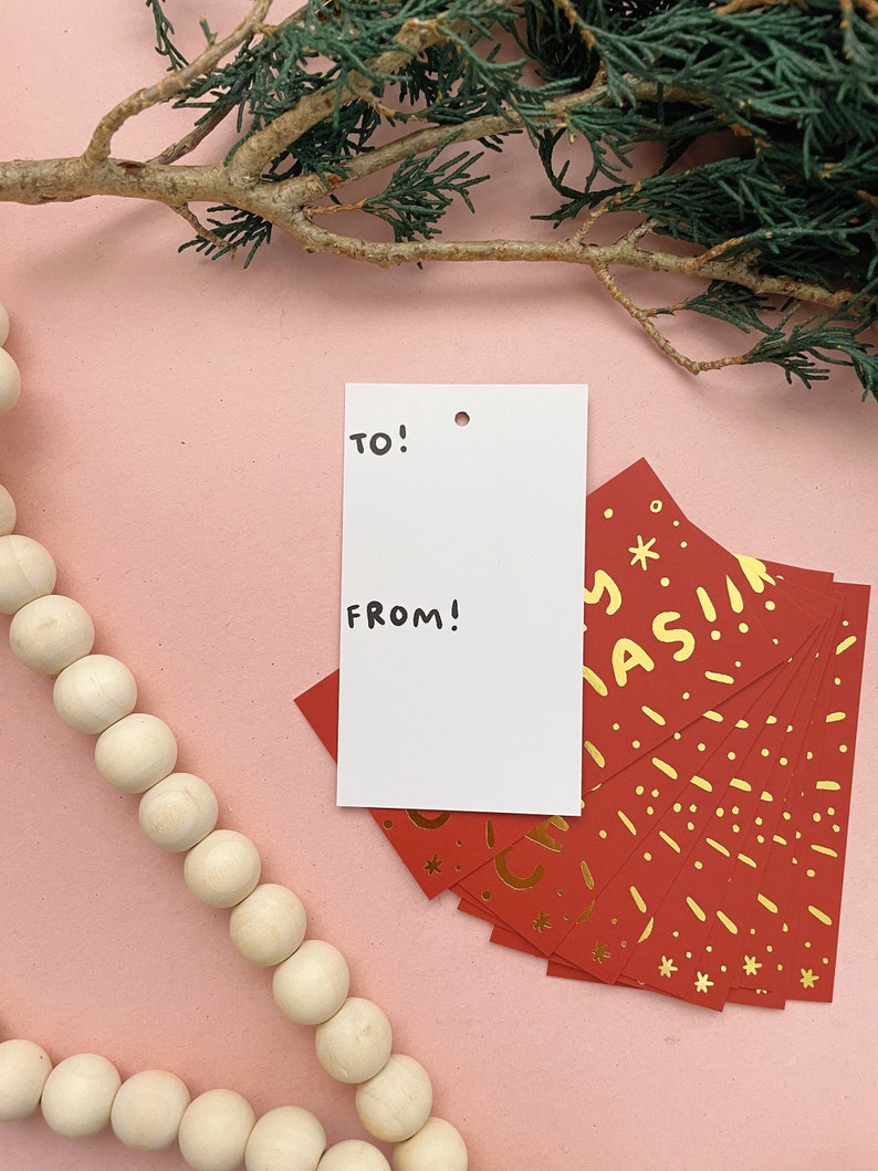 May include: A white gift tag with the words "TO!" and "FROM!" written in black marker. The tag is sitting on a pink surface with a stack of red gift tags with gold foil lettering that says "Merry Christmas!"