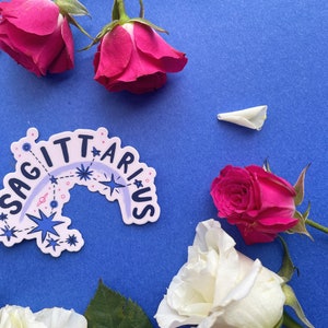 May include: A sticker with the word "Sagittarius" in a rainbow arc with stars and planets on a blue background.  White and pink roses are arranged around the sticker.