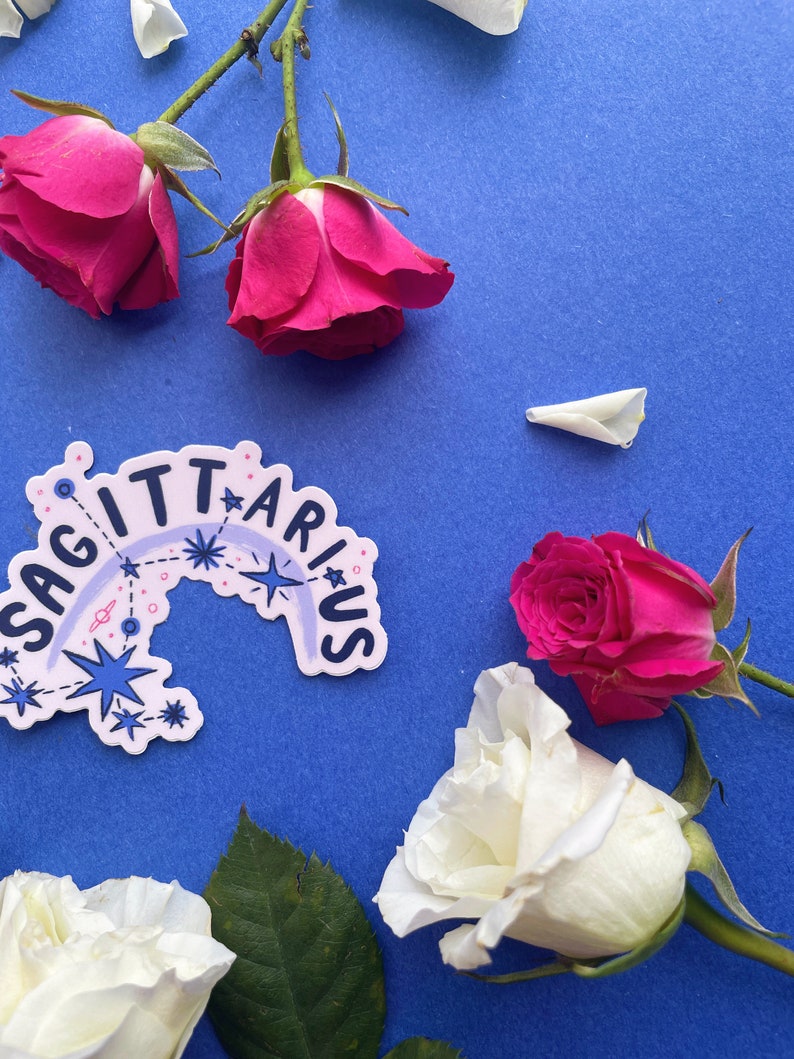 May include: A sticker with the word "Sagittarius" in a rainbow arc with stars and planets on a blue background.  White and pink roses are arranged around the sticker.