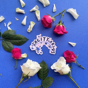 May include: A blue background with white rose petals scattered around a sticker that says "Sagittarius" in a pink and blue font with stars. There are also white and pink roses on the background.
