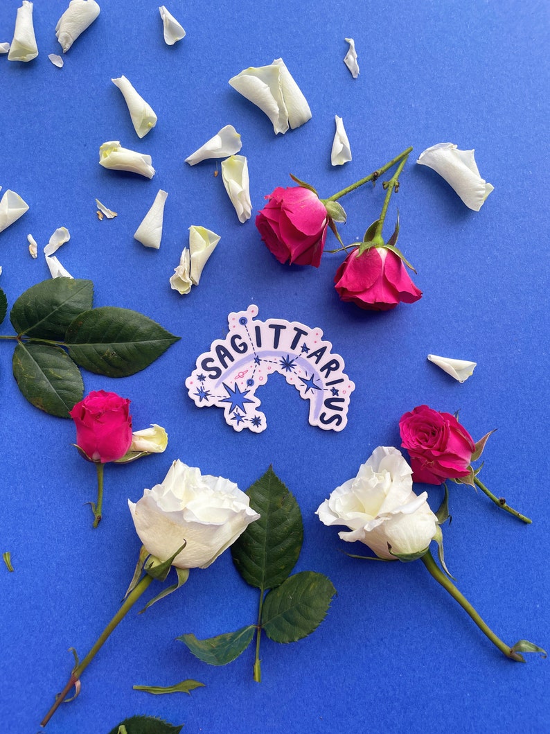 May include: A blue background with white rose petals scattered around a sticker that says "Sagittarius" in a pink and blue font with stars. There are also white and pink roses on the background.