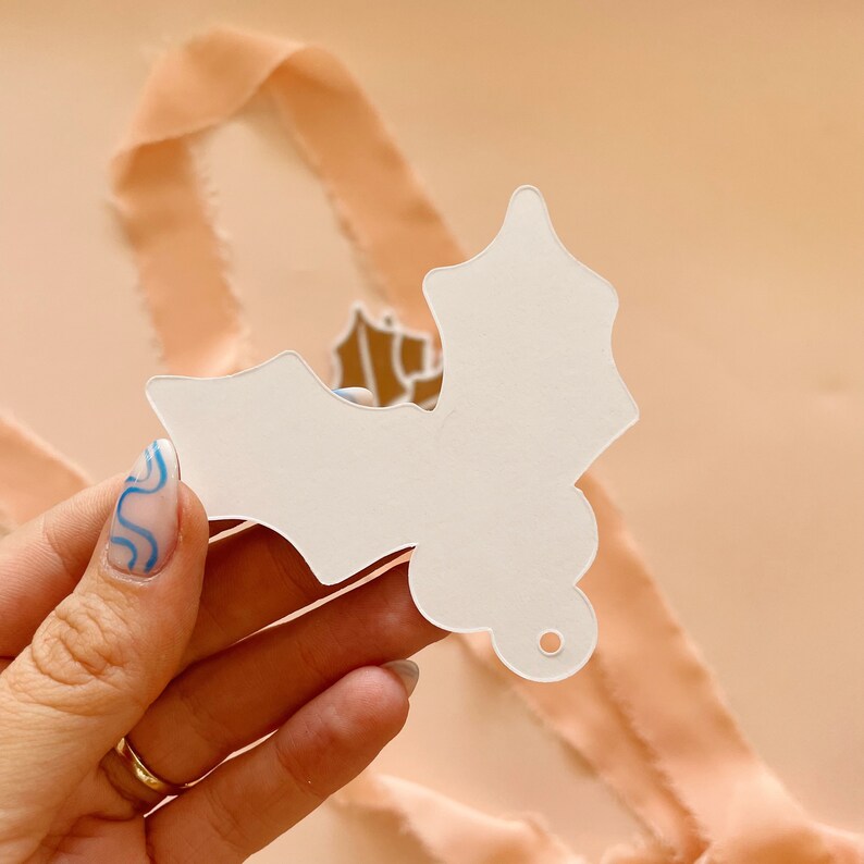 May include: White paper holly leaf ornament with a hole punched at the top for hanging. The ornament is in the shape of a holly leaf with a pointed tip and serrated edges.