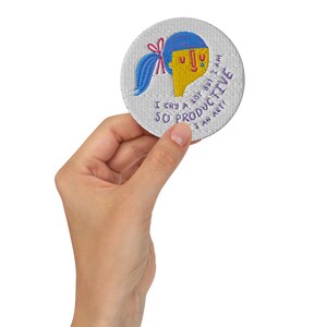 K&ouml;nnte beinhalten: Ein runder gestickter Aufn&auml;her mit einer Cartoon-Illustration einer Person mit blauen Haaren und einem gelben Gesicht. Der Text auf dem Aufn&auml;her lautet "Ich weine viel, aber ich bin so produktiv, stimmt's?"