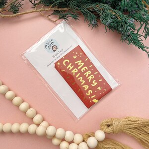 May include: A pack of 10 red gift tags with gold foil lettering that says "Merry Christmas!" The tags are packaged in a clear plastic bag with a white card that says "Abbie Ren 10 Gift Tags Designed in Las Vegas, NV | AbbieRen.com"