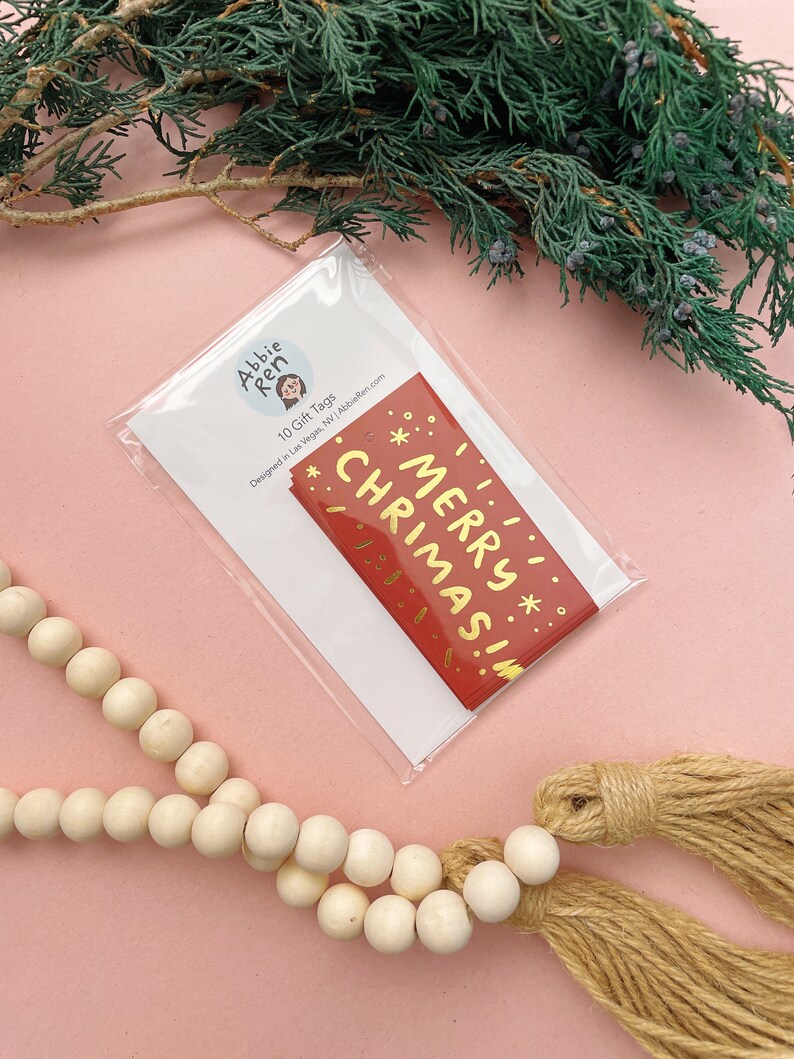 May include: A pack of 10 red gift tags with gold foil lettering that says "Merry Christmas!" The tags are packaged in a clear plastic bag with a white card that says "Abbie Ren 10 Gift Tags Designed in Las Vegas, NV | AbbieRen.com"
