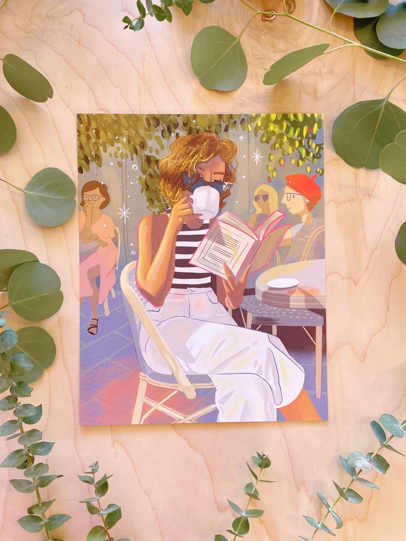 May include: A woman sits at a cafe table, reading a book and drinking from a white mug. She is wearing a striped shirt, white pants, and a black face mask. The cafe is decorated with greenery and has a light blue and pink color scheme.