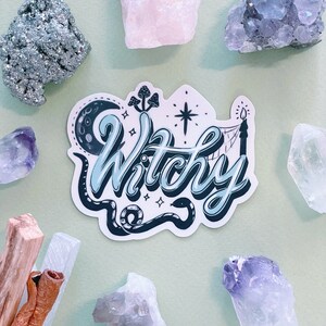 May include: A die-cut sticker featuring the word Witchy in a light blue and dark blue script font. The design includes a crescent moon, a snake, mushrooms, a star, and a candle. The sticker is surrounded by various raw crystals and wood sticks.