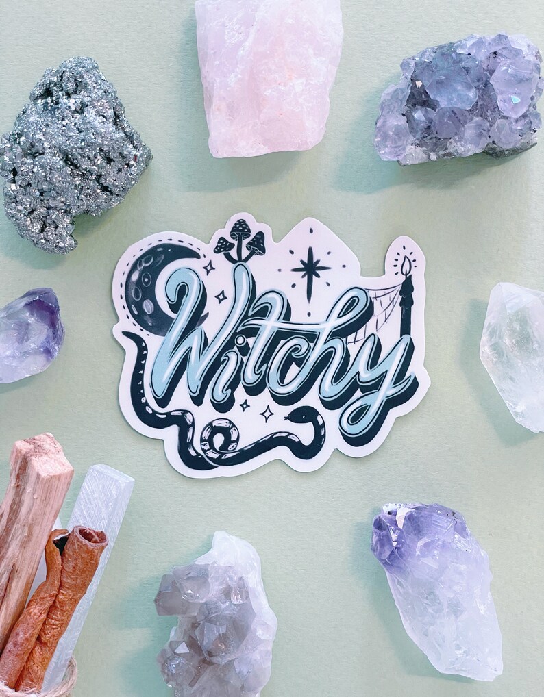 May include: A die-cut sticker featuring the word Witchy in a light blue and dark blue script font. The design includes a crescent moon, a snake, mushrooms, a star, and a candle. The sticker is surrounded by various raw crystals and wood sticks.