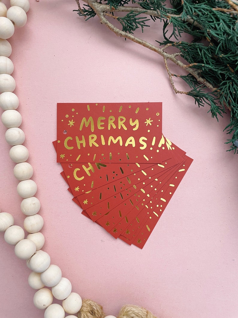 May include: A stack of red gift tags with gold foil lettering that reads "*MERRY* CHRISTMAS!" The tags are decorated with gold foil stars and dots.