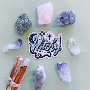 May include: A sticker with the text Witchy in blue and black script, surrounded by a crescent moon, mushrooms, a star, and a snake. The sticker is arranged in a circle with raw crystals, including pyrite, rose quartz, amethyst, and wood sticks.