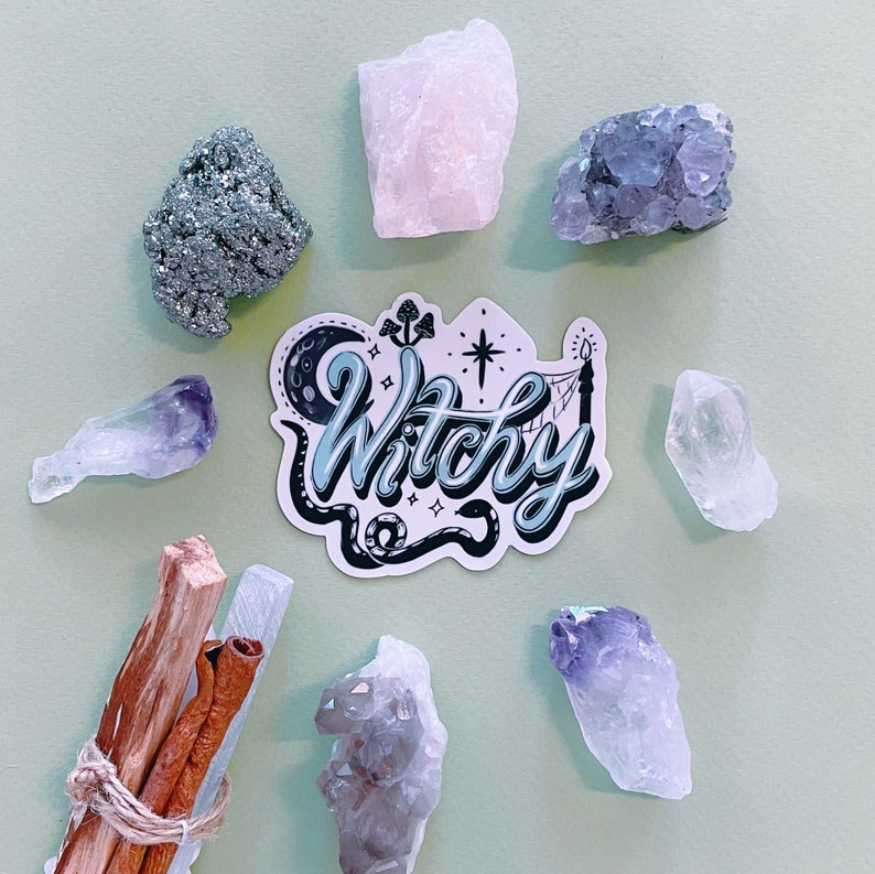 May include: A sticker with the text Witchy in blue and black script, surrounded by a crescent moon, mushrooms, a star, and a snake. The sticker is arranged in a circle with raw crystals, including pyrite, rose quartz, amethyst, and wood sticks.