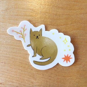 Op de afbeelding: Een gestanste sticker met een gestileerde bruine kat met een klein takje roze bessen. De kat zit met zijn voorpoot omhoog. Gele sterren en een oranje sterrenburst zijn ook in het ontwerp opgenomen.
