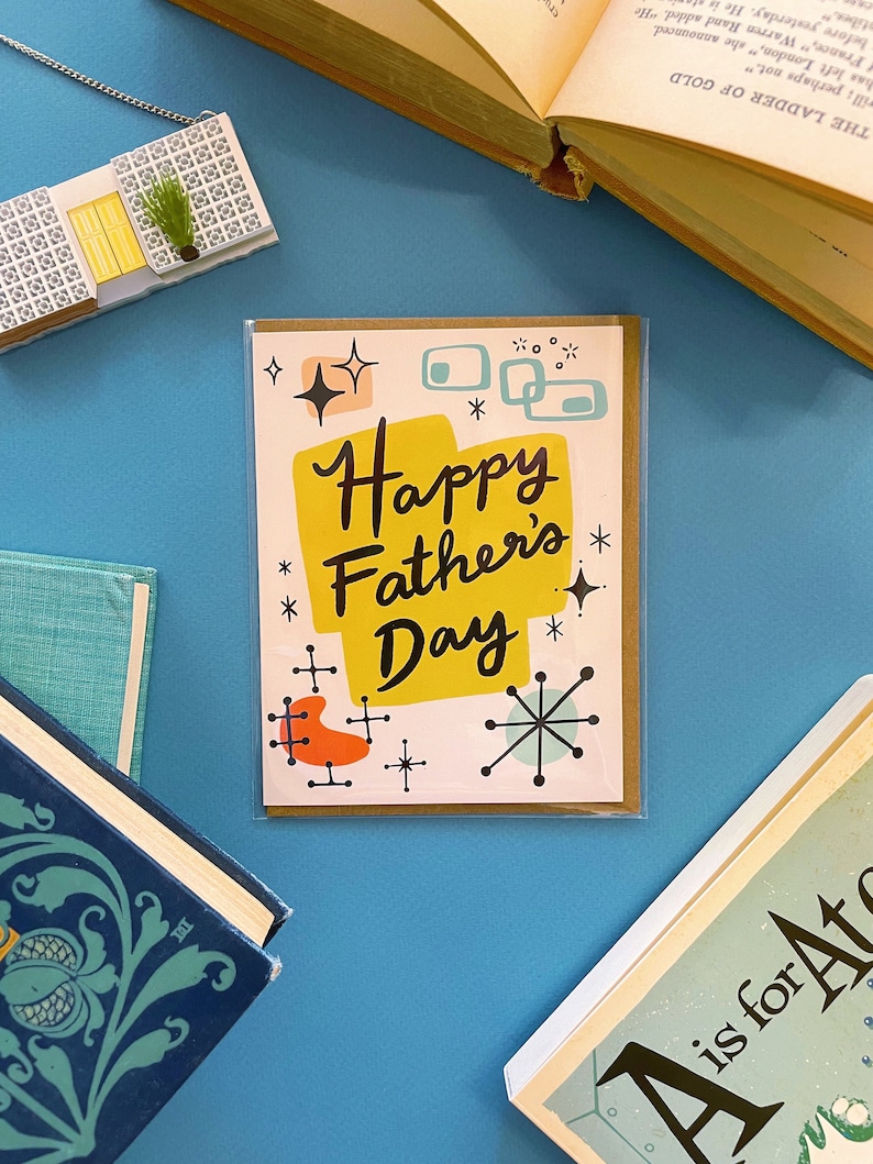 May include: A card with a retro mid-century modern design. The card is yellow, orange, blue, and white with a black and white starburst design. The text on the card reads "Happy Father's Day".
