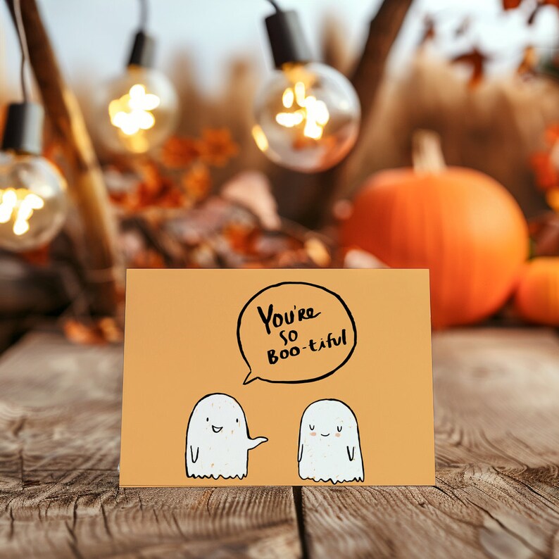 Op de afbeelding: Een lichtoranje kaart met de tekst "You're so Boo-tiful" in zwarte stift. Twee witte geestillustraties staan op de kaart, een met een glimlach en de andere met gesloten ogen. De kaart ligt op een houten oppervlak.