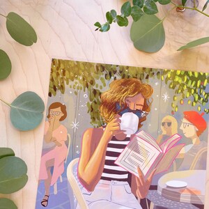 May include: A woman with blonde hair sits at a table in a cafe, reading a book and drinking coffee. She is wearing a striped shirt and white pants. The cafe is decorated with plants and flowers.