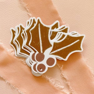 May include: A stack of brown holly leaf gift tags with white outlines and brown berries. The tags are on a peach colored background.