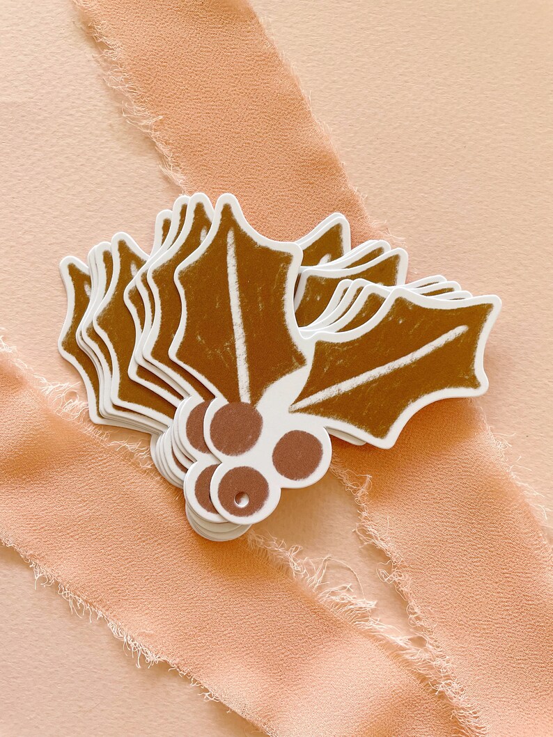 May include: A stack of brown holly leaf gift tags with white outlines and brown berries. The tags are on a peach colored background.