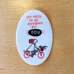 May include: White oval sticker with a black and white illustration of a person riding a bicycle with a basket of flowers. The text on the sticker reads "No need to be anybody but you".