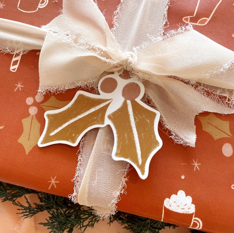 Puede incluir: Una caja de regalo marr&oacute;n con una cinta blanca y un adorno de hoja de acebo marr&oacute;n. La caja est&aacute; envuelta en papel con un patr&oacute;n de copo de nieve blanco.