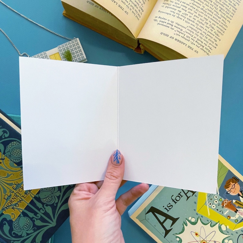 May include: A blank white card with a folded edge, held open by a hand with blue nail polish. The card is on a blue surface with a book on the left and a book on the right. The book on the right has a blue cover with the text "A is for" and an illustration of an atom.