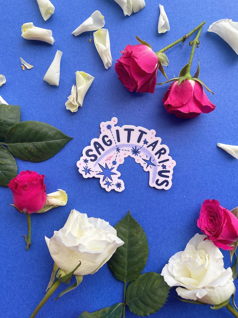 May include: A sticker with the word "Sagittarius" in a rainbow arc with stars and a constellation design. The sticker is on a blue background with white rose petals and pink roses.