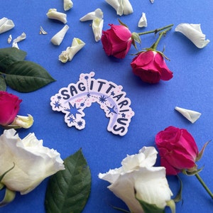 May include: A white sticker with a blue constellation design and the text "SAGITTARIUS" on a blue background with white and pink roses.