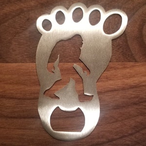 Può includere: Un apribottiglie in metallo argentato a forma di impronta di piede con all'interno la silhouette di Bigfoot.