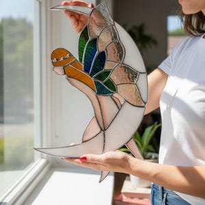 May include: A stained glass sculpture in the shape of a crescent moon with a stylized bird or butterfly design. The artwork features white, orange, green, and iridescent glass pieces, held together by dark metal lines.
