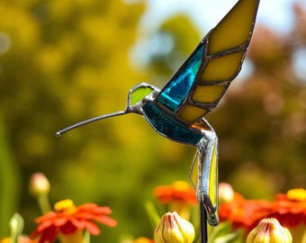 Può includere: Un paletto da giardino a colibrì in vetro colorato con pannelli di vetro giallo, blu e verde. L'uccello è in volo, con il becco lungo esteso. Il paletto è in un giardino con fiori arancioni.