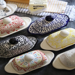 May include: Six ceramic butter dishes with different designs. The dishes are white with blue, black, red, yellow, and multi-colored designs. The dishes have a domed lid with a knob on top.