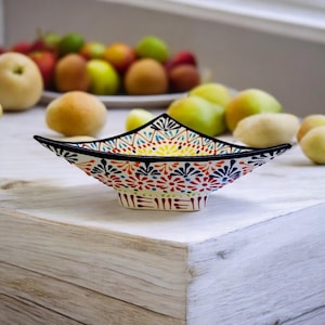 May include: A white ceramic bowl with a colourful geometric design. The bowl is shaped like a star and has a wide, shallow base. The design features red, blue, yellow, and green colours.