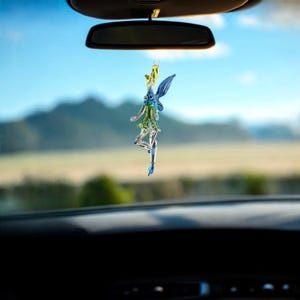 以下が含まれることがあります： 車の後部座席のミラーに、青と緑の羽根を持つガラス製の妖精の置物がぶら下がっています。妖精は緑色のドレスを着ており、髪には黄色の花を付けています。
