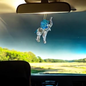 May include: A clear crystal elephant figurine with blue accents hanging from a rearview mirror in a car. The elephant is facing forward with its trunk raised.