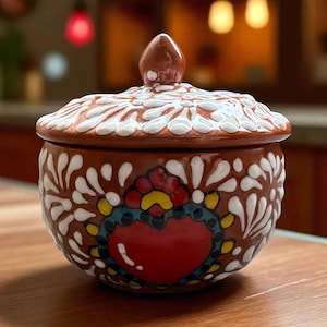 May include: A brown ceramic jar with a lid, adorned with white floral patterns and a central heart design. The heart is red with blue, yellow, and green accents. The jar is likely handmade and suitable for kitchen or home decor.